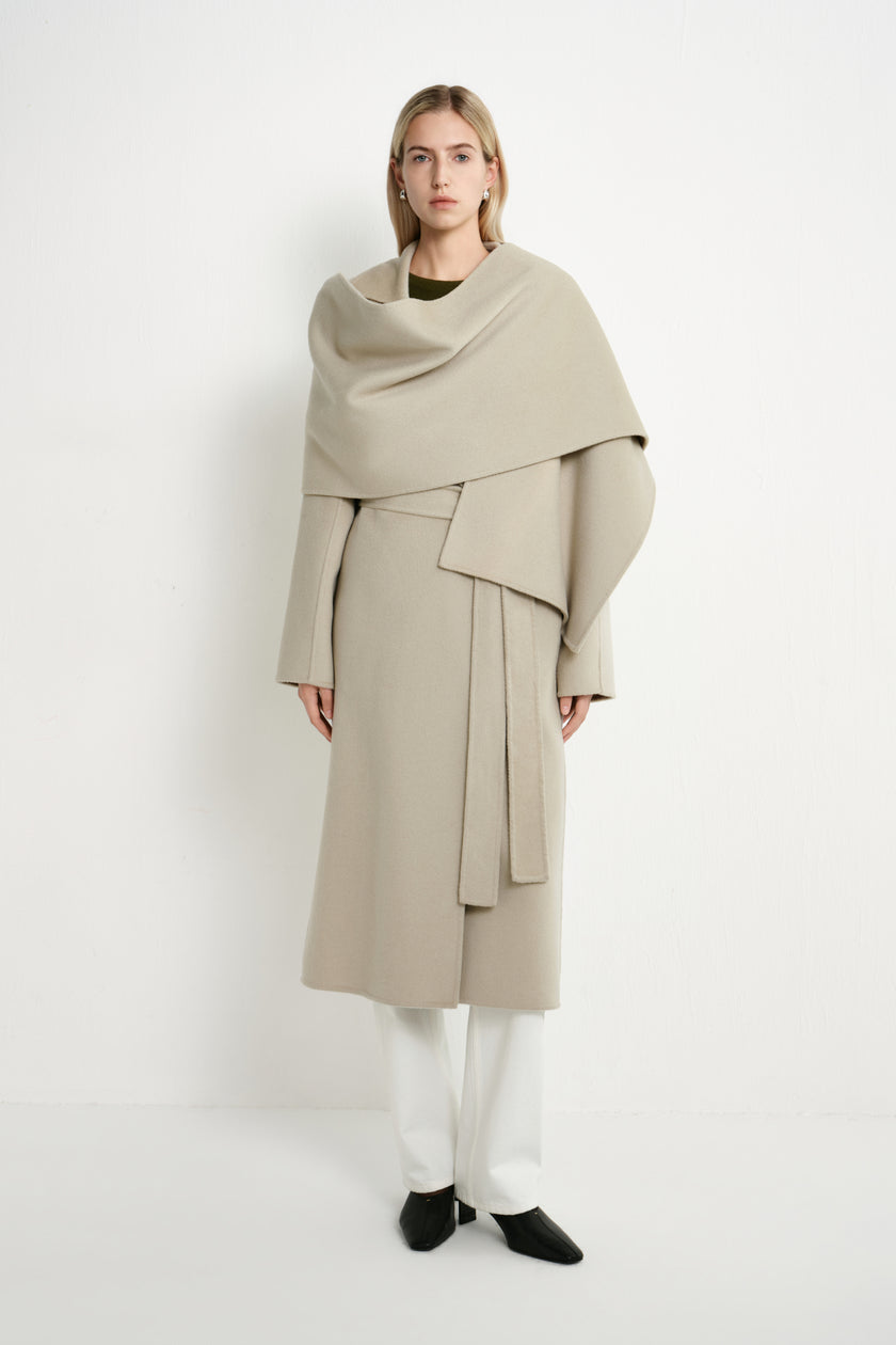 A model stands against a plain white studio background wearing a long, draped beige coat with a wide shawl-like collar, white pants, and black shoes.