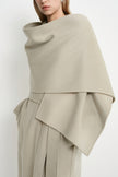 A close-up of a model wearing a draped, wide shawl-style wrap or poncho in a light warm gray-beige tone, paired with matching draped trousers against a plain light background.