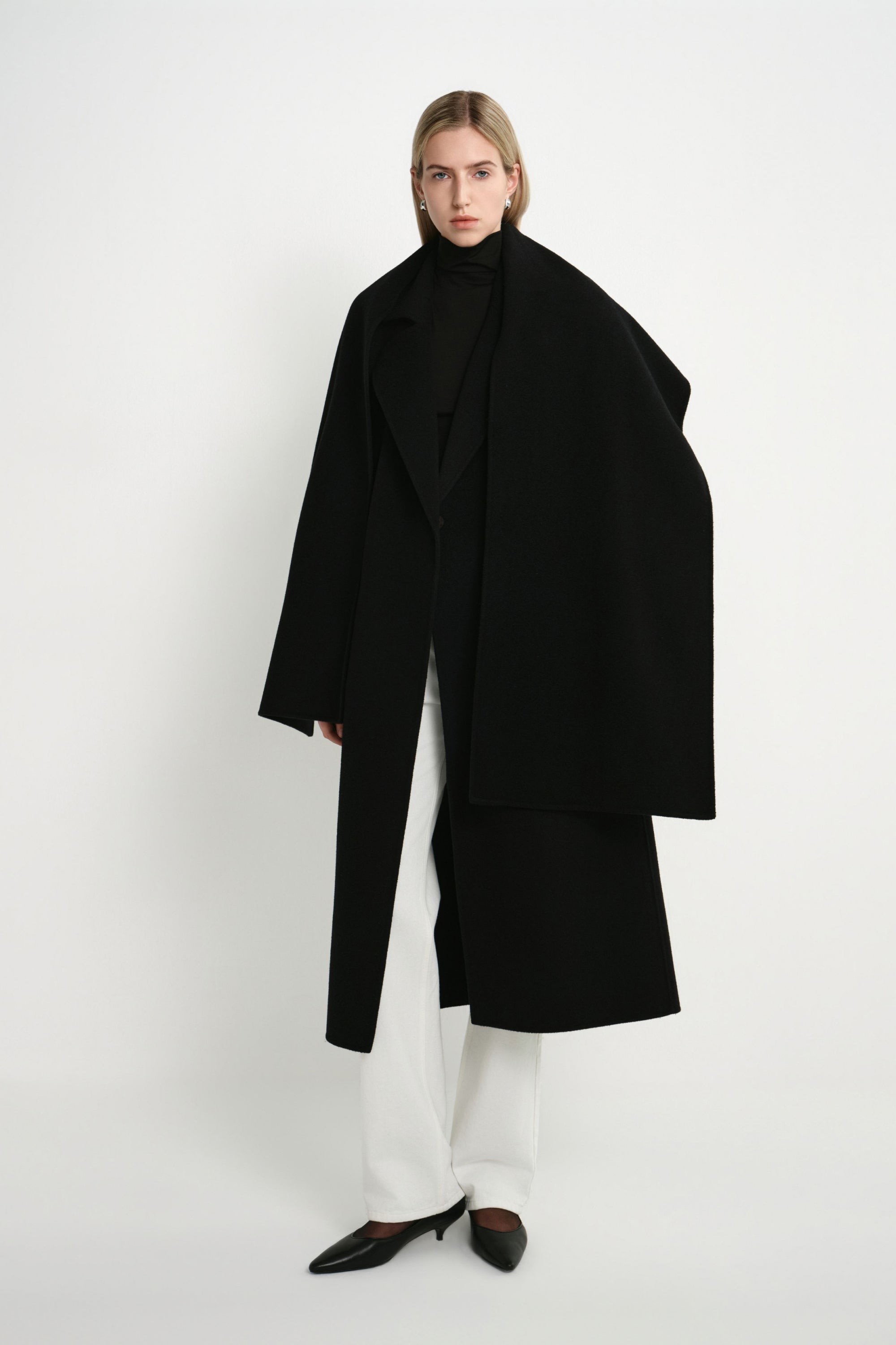 A model wearing the Rue Sophie Grand Scarf Coat in black with an oversized scarf draped over the shoulders, long sleeves, and a relaxed midi silhouette, full-length front view.