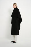 A model wearing the Rue Sophie Grand Scarf Coat in black, highlighting the asymmetric scarf drape, belted waist, and long straight silhouette, three-quarter side view.