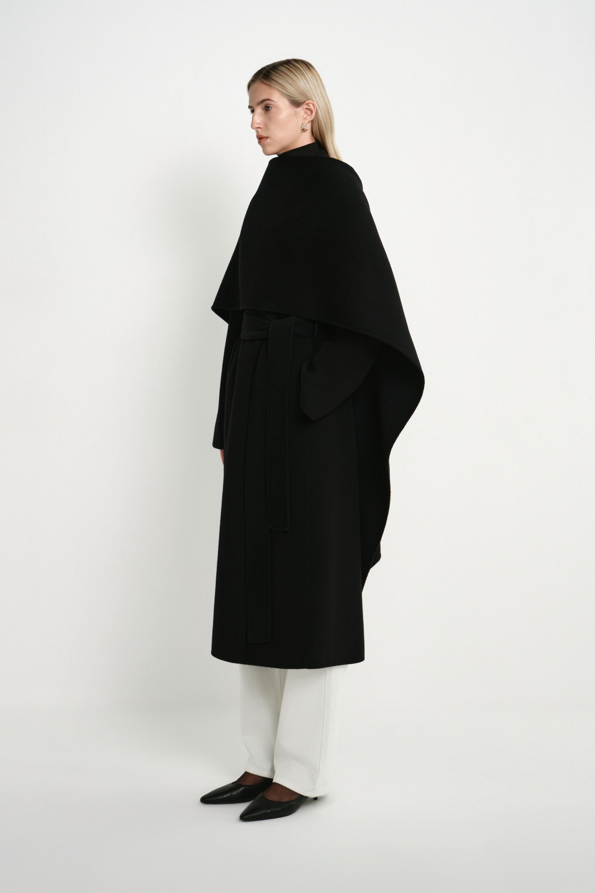 A model wearing the Rue Sophie Grand Scarf Coat in black, highlighting the asymmetric scarf drape, belted waist, and long straight silhouette, three-quarter side view.