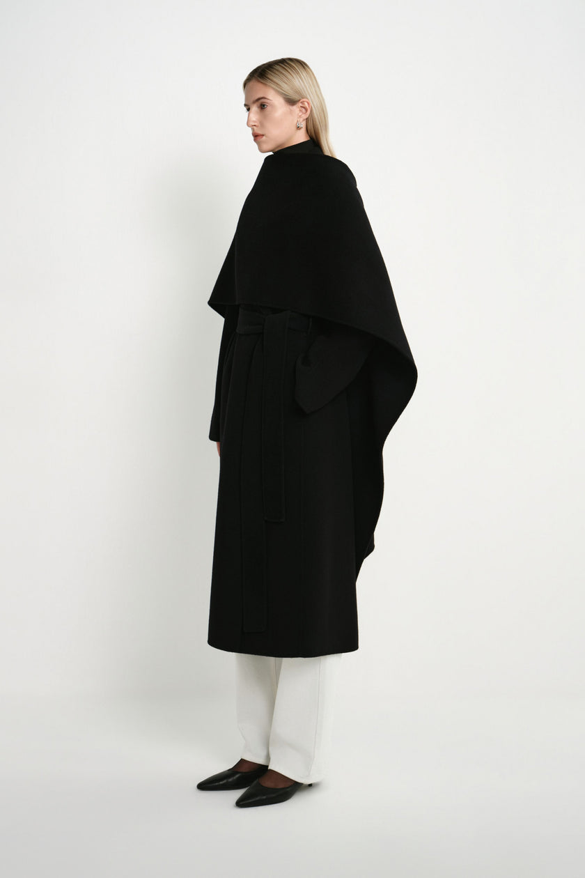 A model wearing the Rue Sophie Grand Scarf Coat in black, highlighting the asymmetric scarf drape, belted waist, and long straight silhouette, three-quarter side view.