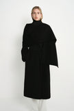 A model wearing the Rue Sophie Grand Scarf Coat in black with a wrap-style scarf panel, self-tie waist belt, and structured midi-length silhouette, front view.