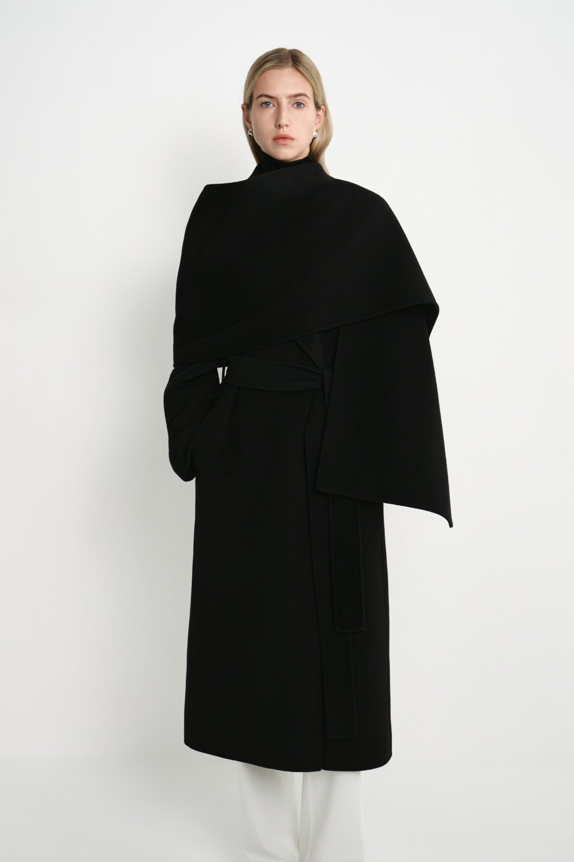 A model wearing the Rue Sophie Grand Scarf Coat in black with a wrap-style scarf panel, self-tie waist belt, and structured midi-length silhouette, front view.