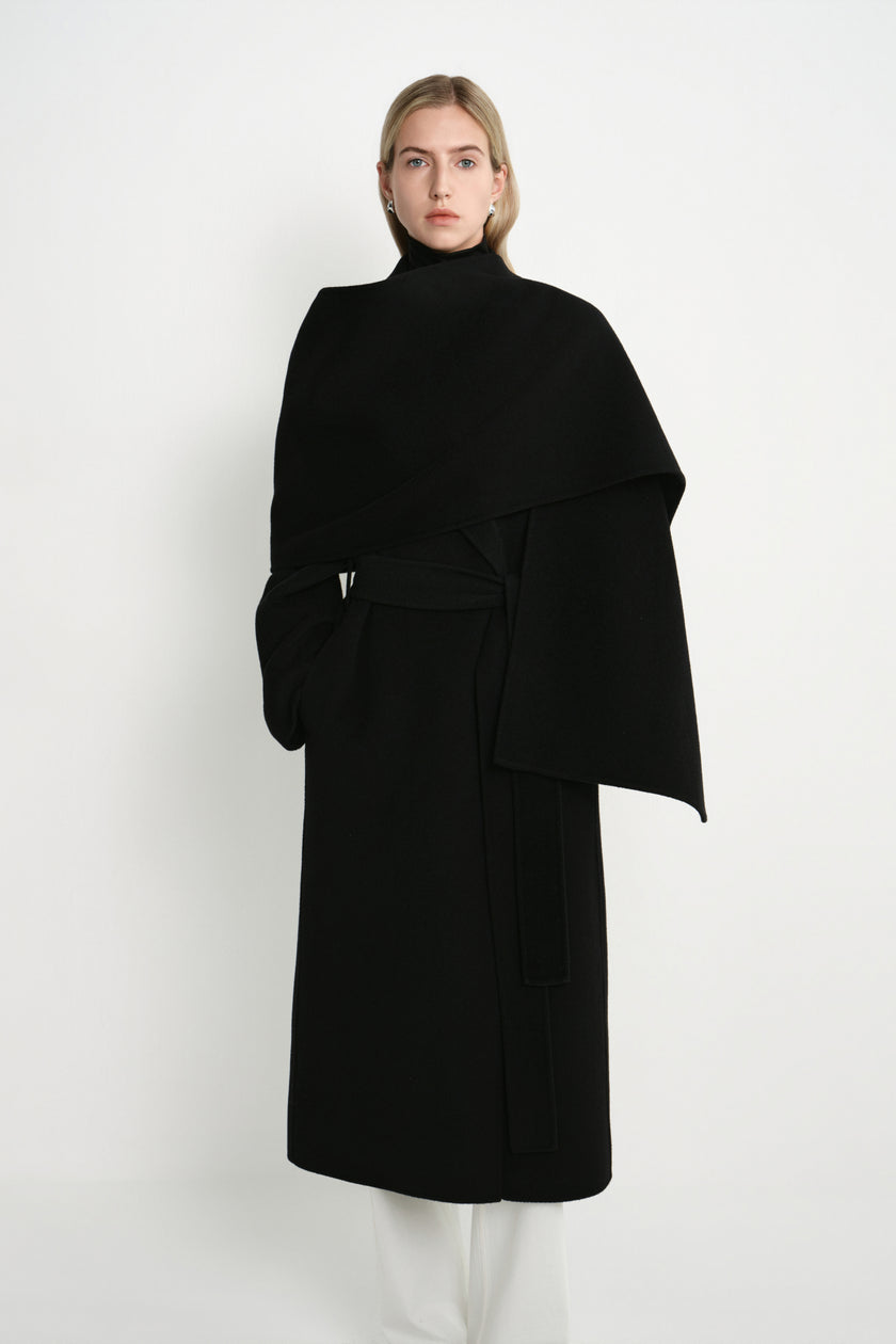 A model wearing the Rue Sophie Grand Scarf Coat in black with a wrap-style scarf panel, self-tie waist belt, and structured midi-length silhouette, front view.