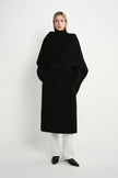 A model wearing the Rue Sophie Grand Scarf Coat in black, featuring an oversized draped scarf panel, belted waist, long sleeves, and a clean midi silhouette, front view.