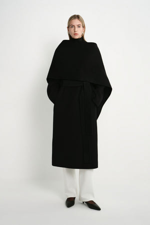 A model wearing the Rue Sophie Grand Scarf Coat in black, featuring an oversized draped scarf panel, belted waist, long sleeves, and a clean midi silhouette, front view.