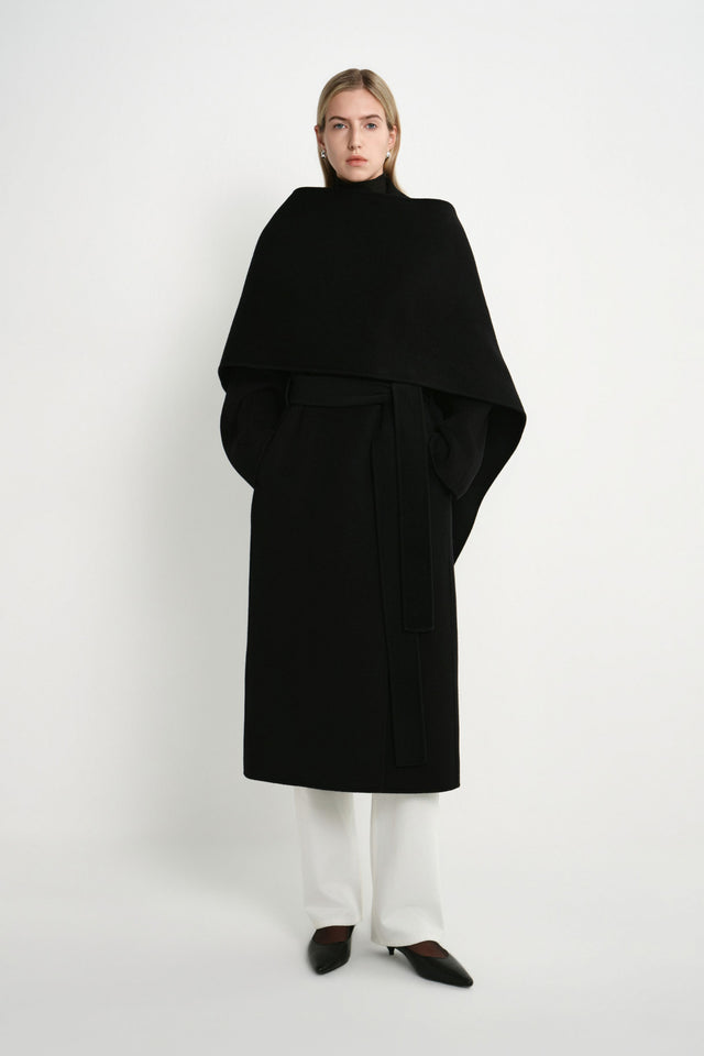 A model wearing the Rue Sophie Grand Scarf Coat in black, featuring an oversized draped scarf panel, belted waist, long sleeves, and a clean midi silhouette, front view.