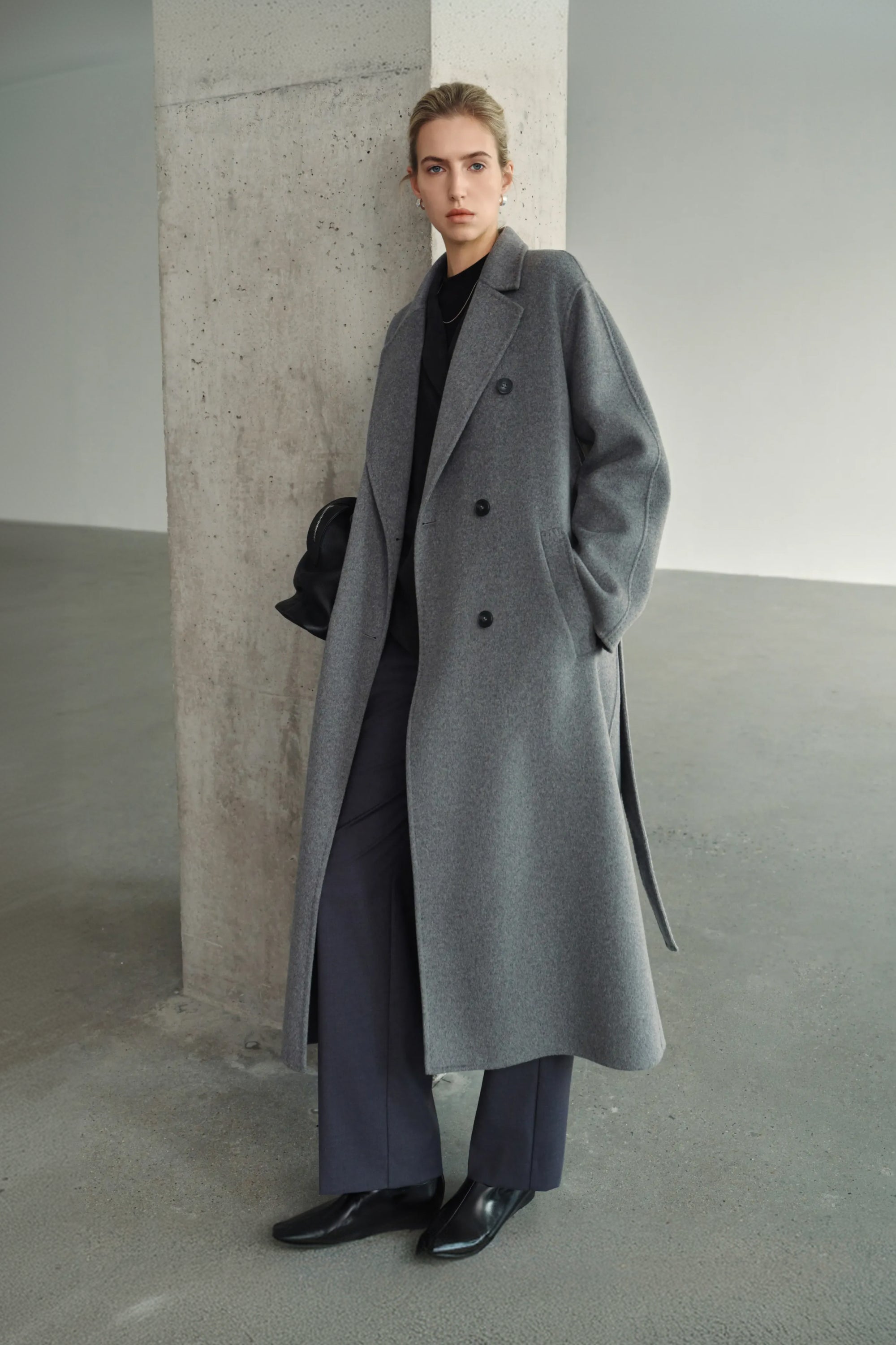 A model wearing the Rue Sophie Martine Belted Coat in grey, featuring wide notched lapels, side pockets, and a relaxed ankle-length silhouette, three-quarter front view.