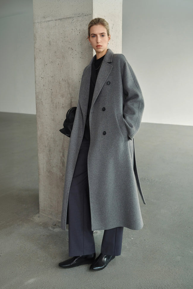 A model wearing the Rue Sophie Martine Belted Coat in grey, featuring wide notched lapels, side pockets, and a relaxed ankle-length silhouette, three-quarter front view.