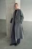 A model wearing the Rue Sophie Martine Belted Coat in grey, featuring wide notched lapels, side pockets, and a relaxed ankle-length silhouette, three-quarter front view.