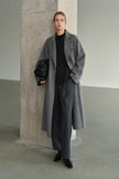 A model wearing the Rue Sophie Martine Belted Coat in grey with an oversized longline shape, wide lapels, button-front closure, and relaxed sleeves, three-quarter front view.