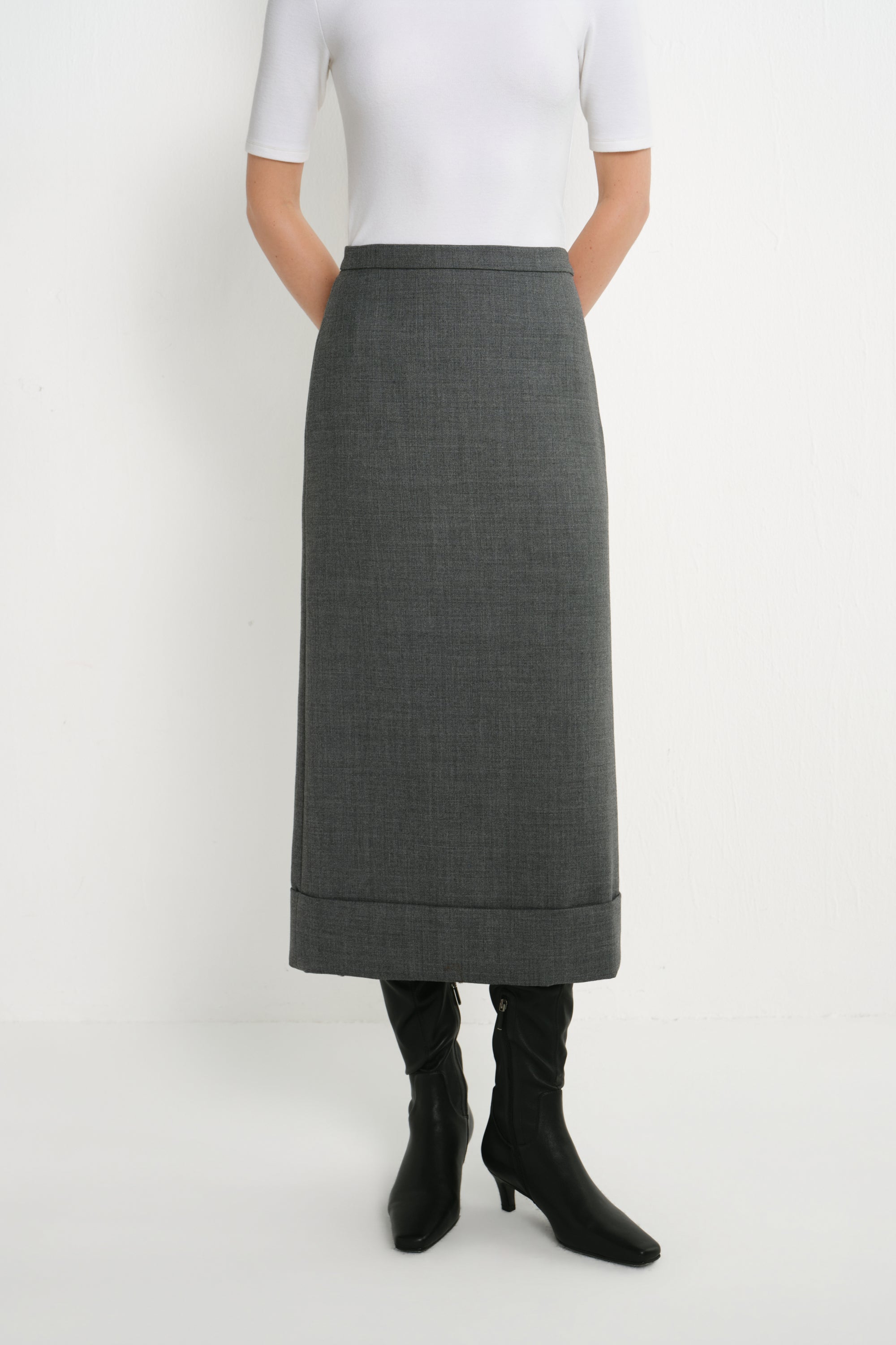 A model wearing a dark gray, knee-length textured skirt with rolled cuffs, paired with a white short-sleeve top and black knee-high boots, shot against a plain white studio background.