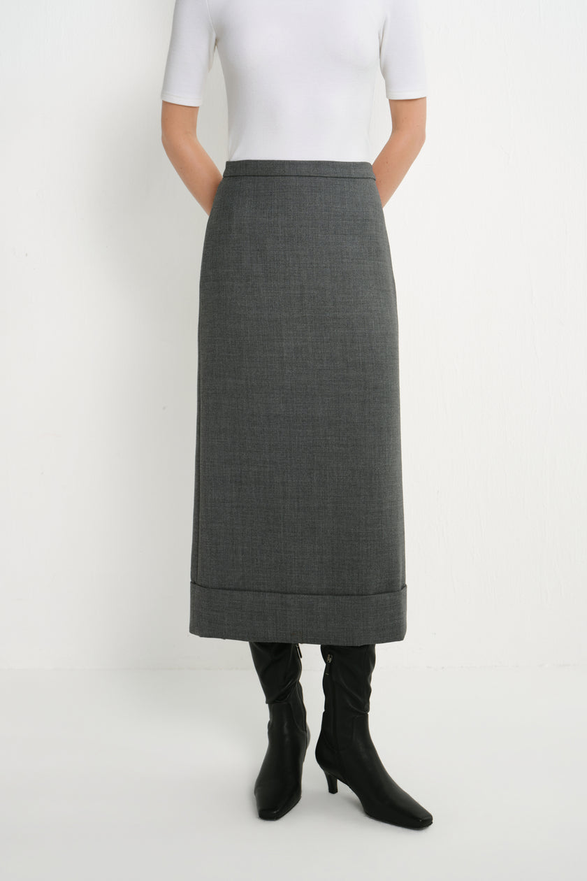 A model wearing a dark gray, knee-length textured skirt with rolled cuffs, paired with a white short-sleeve top and black knee-high boots, shot against a plain white studio background.