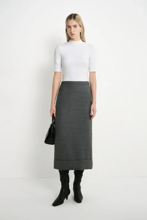 A woman stands against a plain white studio background wearing a white short-sleeve mock-neck top, a dark gray knee-length skirt, black knee-high boots, and holding a black handbag.