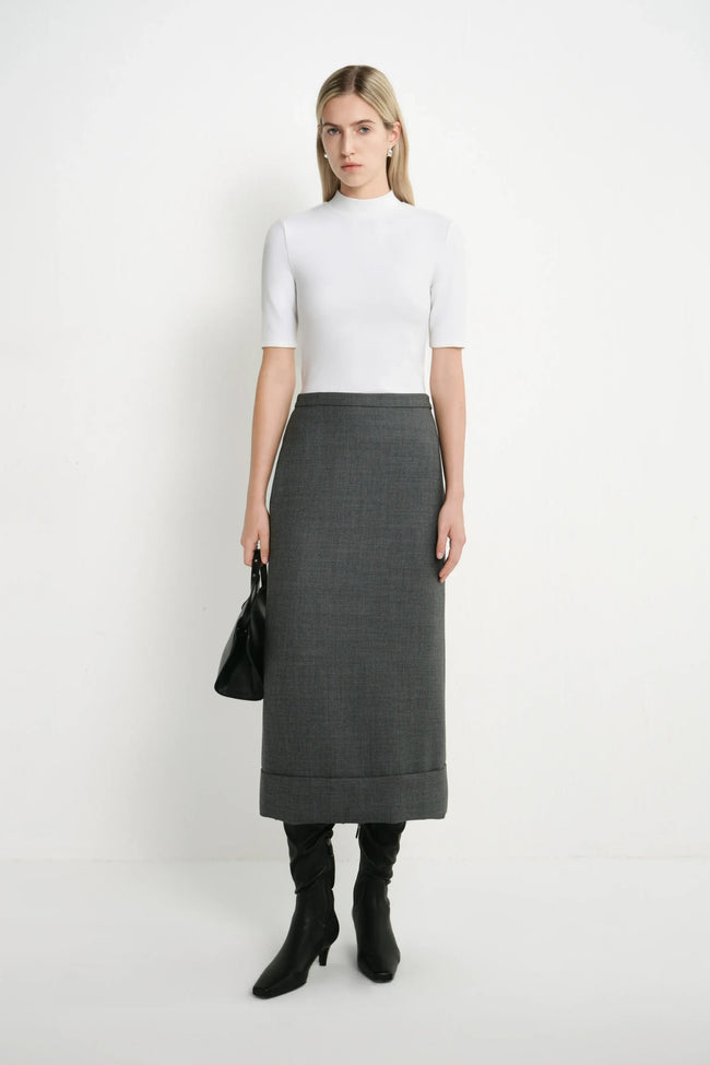 A woman stands against a plain white studio background wearing a white short-sleeve mock-neck top, a dark gray knee-length skirt, black knee-high boots, and holding a black handbag.