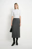 A woman stands against a plain white studio background wearing a white short-sleeve mock-neck top, a dark gray knee-length skirt, black knee-high boots, and holding a black handbag.