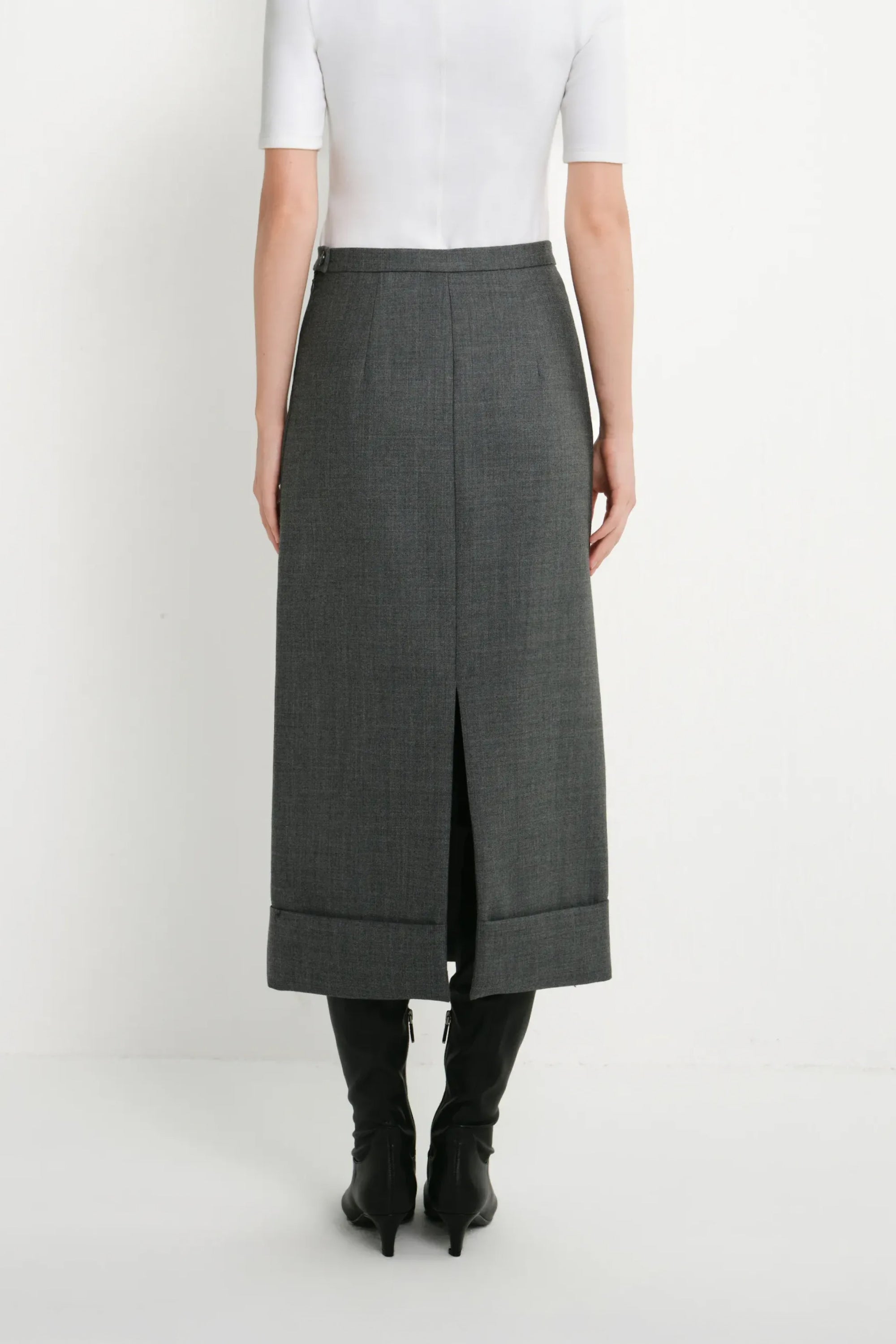 A model from the back wearing a dark gray textured midi skirt with a front slit, styled with a white short-sleeve top and black knee-high boots against a plain white studio background.
