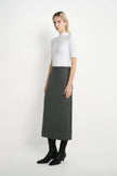 A woman stands in a studio wearing a white short-sleeve mock-neck top, a dark gray midi skirt, and black ankle boots against a plain light background.
