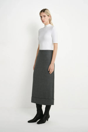 A woman stands in a studio wearing a white short-sleeve mock-neck top, a dark gray midi skirt, and black ankle boots against a plain light background.