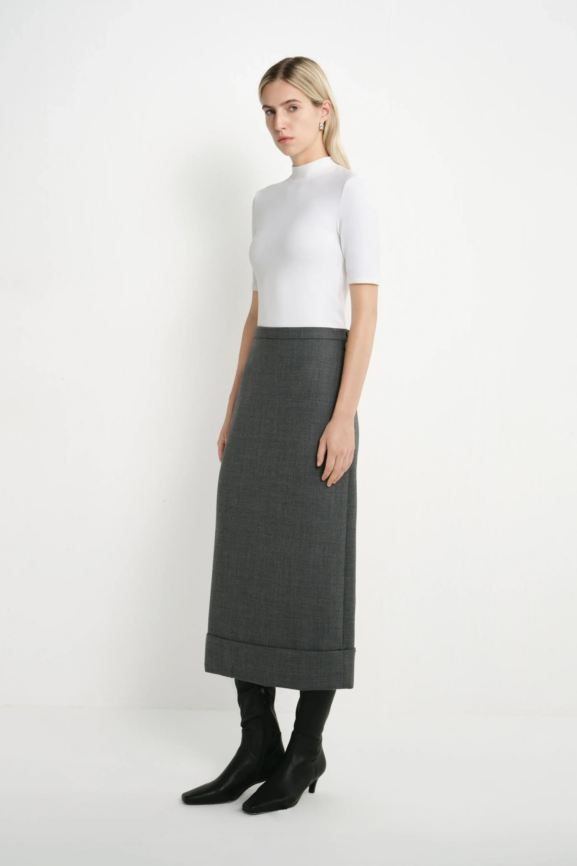 A woman stands in a studio wearing a white short-sleeve mock-neck top, a dark gray midi skirt, and black ankle boots against a plain light background.