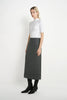 A woman stands in a studio wearing a white short-sleeve mock-neck top, a dark gray midi skirt, and black ankle boots against a plain light background.