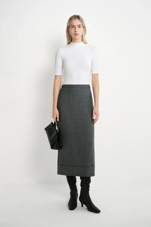 A model stands against a plain white studio background wearing a white short-sleeve mock-neck top, a dark gray textured midi skirt, black knee-high boots, and holding a black clutch bag.