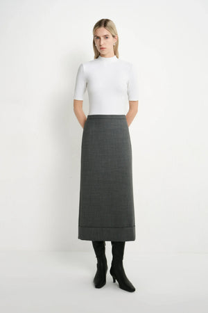 A woman stands against a plain white studio background wearing a white short-sleeve mock-neck top, a dark gray knee-length skirt, and black ankle boots.