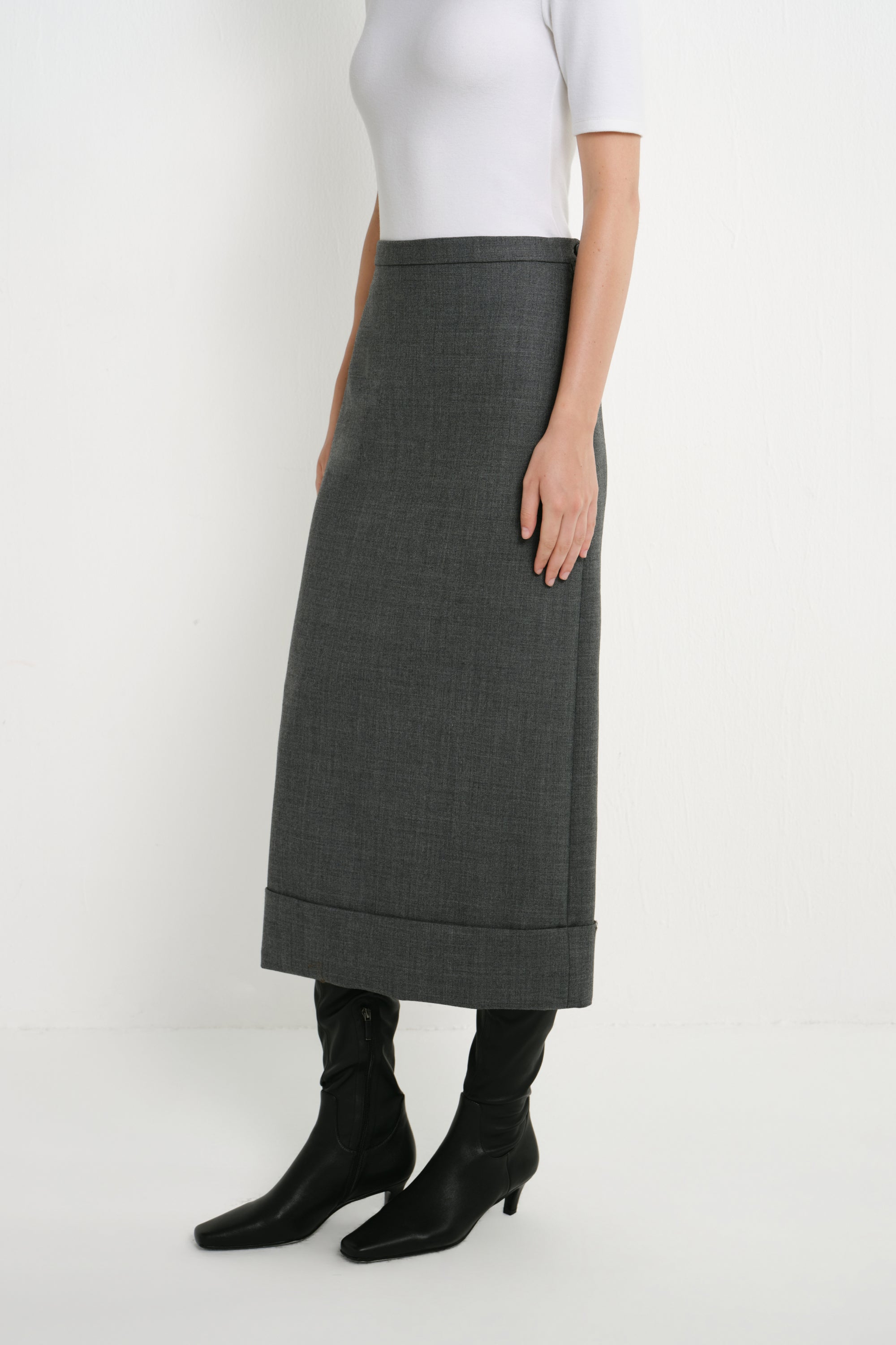A model wearing a dark gray, mid-calf length skirt with rolled cuffs, styled with a white top and black pointed-toe ankle boots against a plain white studio background.
