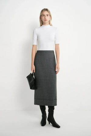 A woman stands against a plain white studio background wearing a fitted white short-sleeve mock-neck top, a dark gray knee-length skirt, black knee-high socks, and black heeled boots, while holding a black clutch.