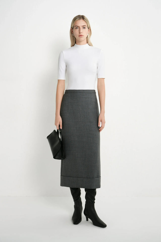 A woman stands against a plain white studio background wearing a fitted white short-sleeve mock-neck top, a dark gray knee-length skirt, black knee-high socks, and black heeled boots, while holding a black clutch.