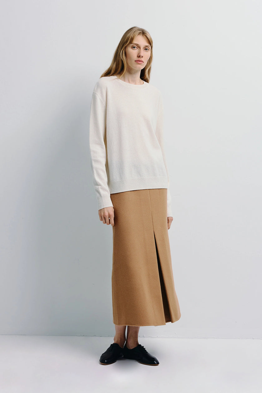 Woman wearing Ambre Pleat Front Midi Skirt in camel, paired with a cream sweater, and black shoes, tailored A-line silhouette
