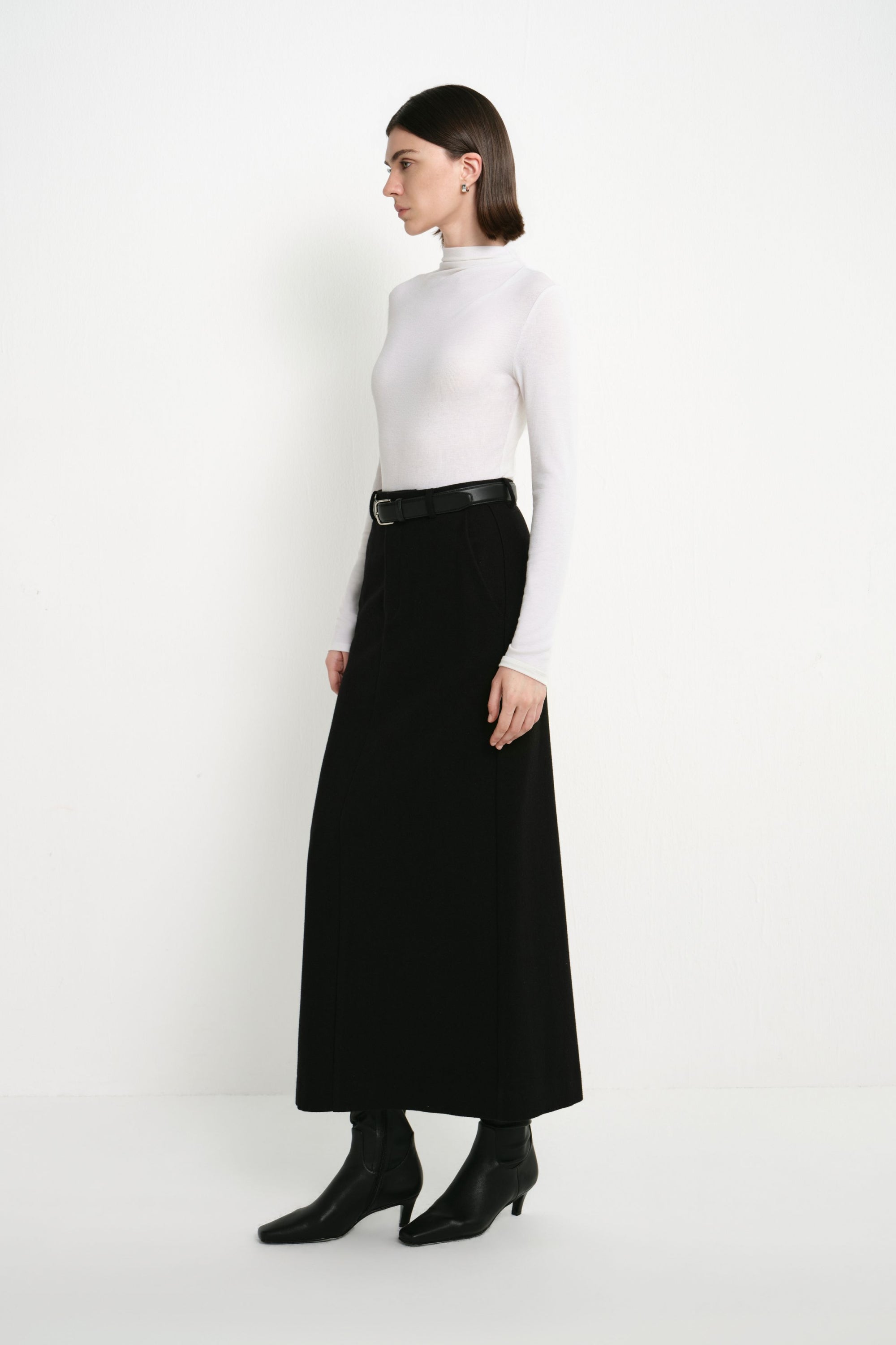 Side view of a model wearing the Rue Sophie Camberly Midi Skirt in black, featuring a high-waisted fit and clean elongated A-line silhouette.