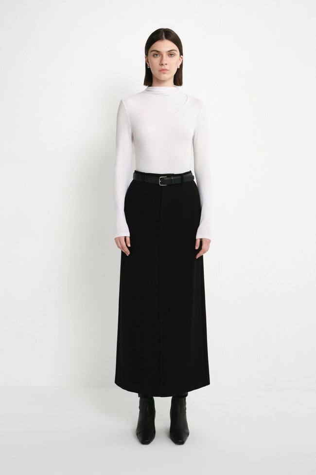 A model stands against a plain white studio background wearing a white mock-neck long-sleeve top, a black midi skirt with a black belt, and black ankle boots.