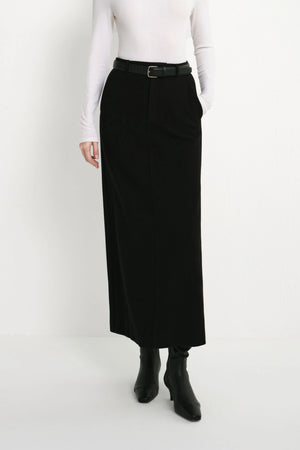 A model wearing a long black skirt with a black belt, paired with a white long-sleeve top and black ankle boots, shown from the waist down against a plain white studio background.