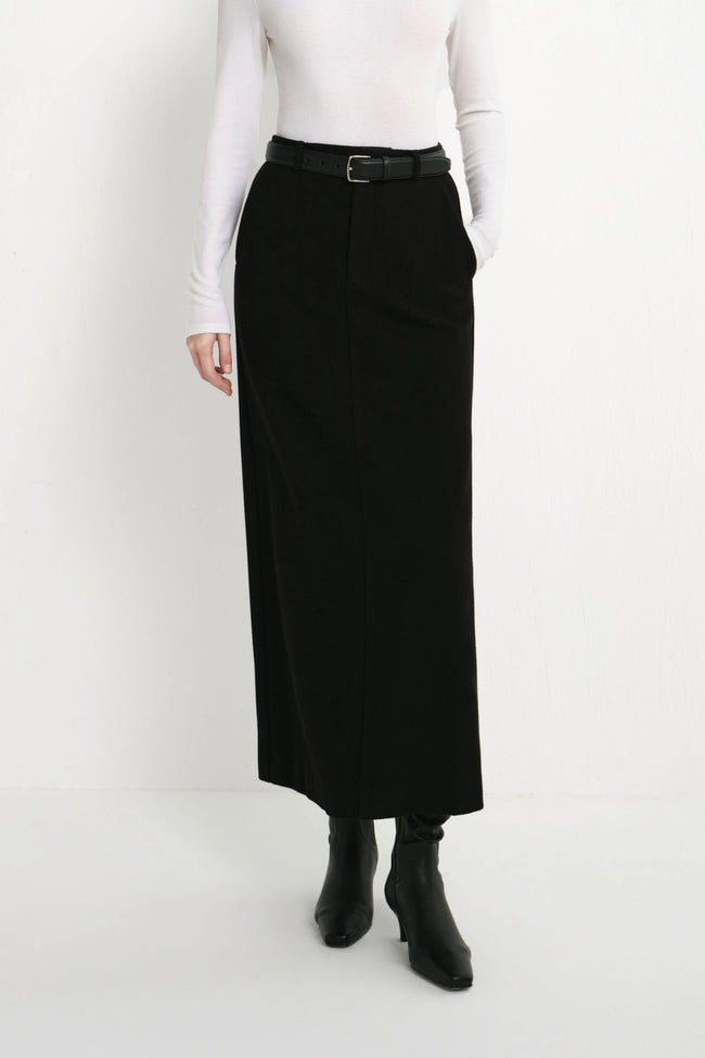 A model wearing a long black skirt with a black belt, paired with a white long-sleeve top and black ankle boots, shown from the waist down against a plain white studio background.