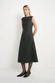 A woman modeling a sleeveless, knee-length dark gray dress with a high round neckline, paired with black tights and black pointed flats, against a plain white studio background.