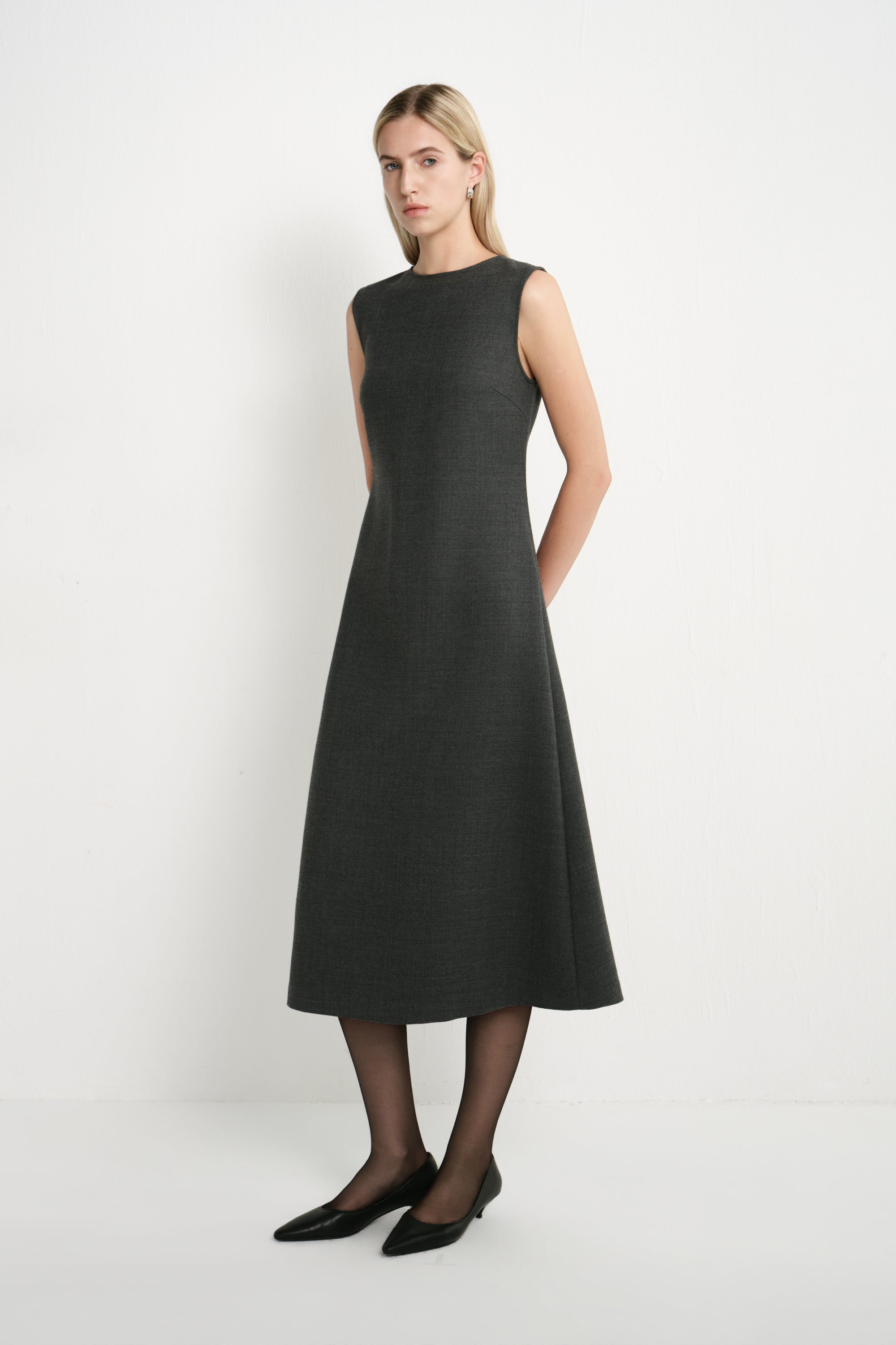 A woman modeling a sleeveless, knee-length dark gray dress with a high round neckline, paired with black tights and black pointed flats, against a plain white studio background.