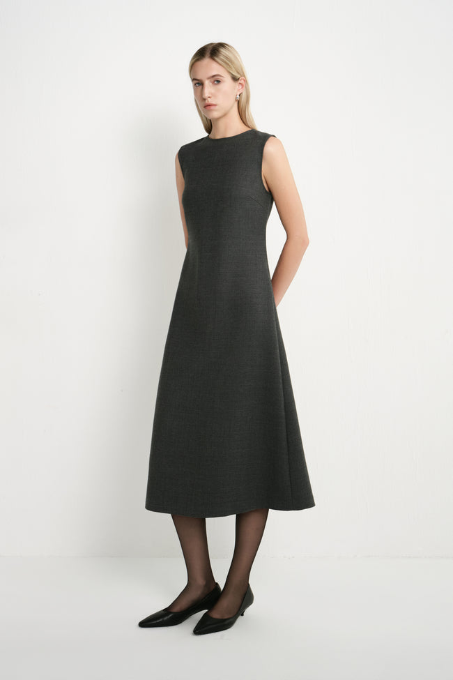 A woman modeling a sleeveless, knee-length dark gray dress with a high round neckline, paired with black tights and black pointed flats, against a plain white studio background.