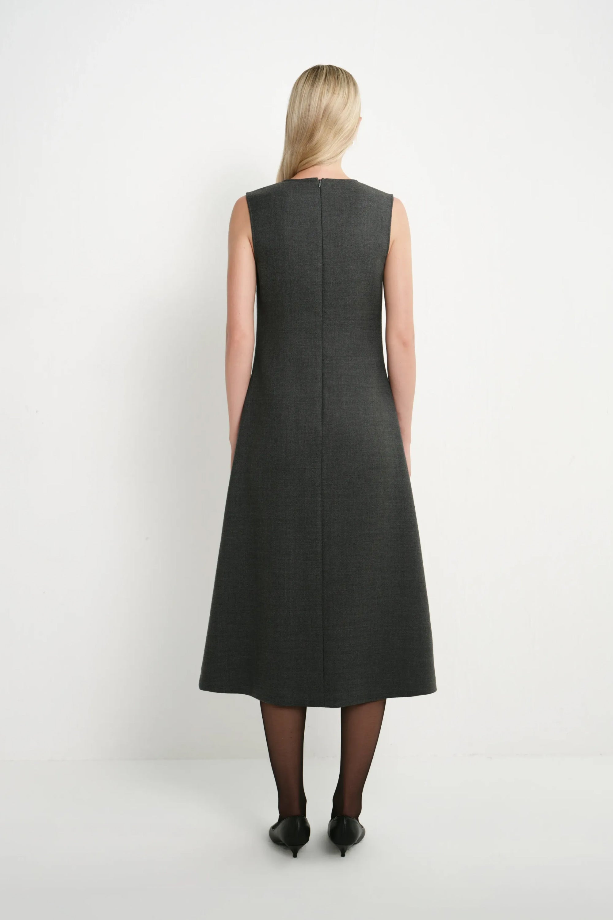 A woman viewed from the back wearing a sleeveless dark gray A-line midi dress, black tights, and black heels against a plain white studio background.