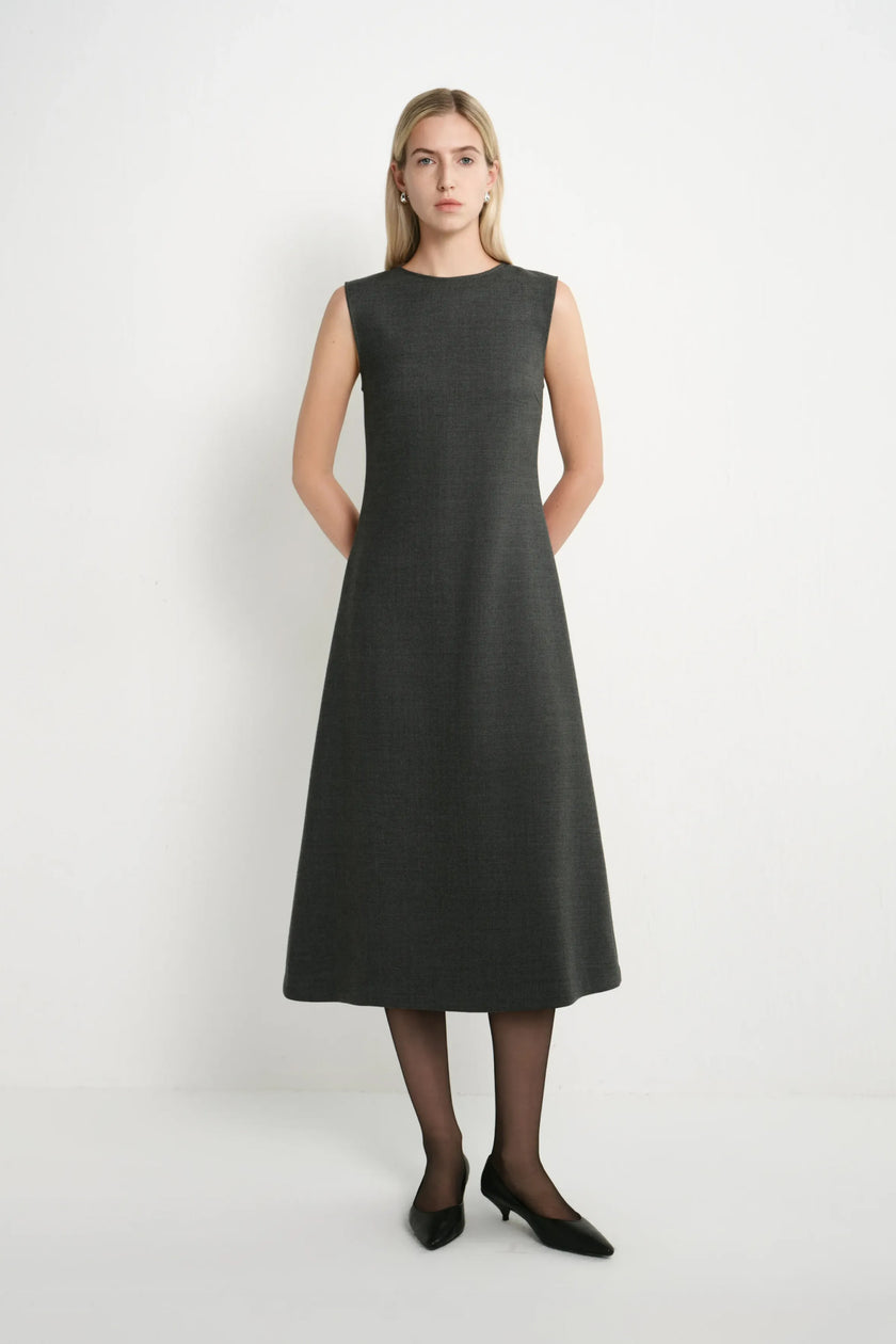 A woman modeling a sleeveless, knee-length dark gray A-line dress with a textured weave, paired with black tights and black pointed-toe heels, against a plain white studio background.