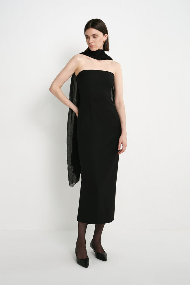A model wearing the Rue Sophie Webster Tube Dress in black with a straight strapless neckline and fitted column silhouette, styled with a sheer black scarf, three-quarter front view.