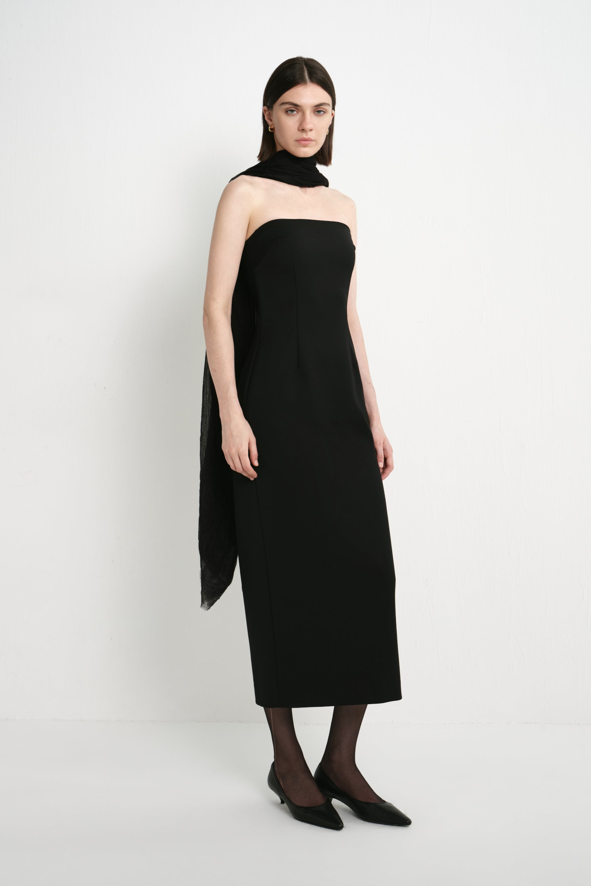 A model wearing the Rue Sophie Webster Tube Dress in black, featuring a strapless neckline, sculpted waist, and streamlined column shape, styled with a sheer black scarf.