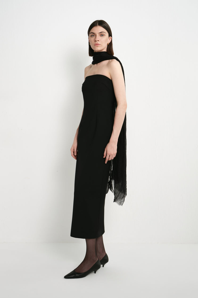 A model wearing the Rue Sophie Webster Tube Dress in black with a straight strapless neckline and fitted mid-calf silhouette, styled with a draped black scarf, three-quarter side view.