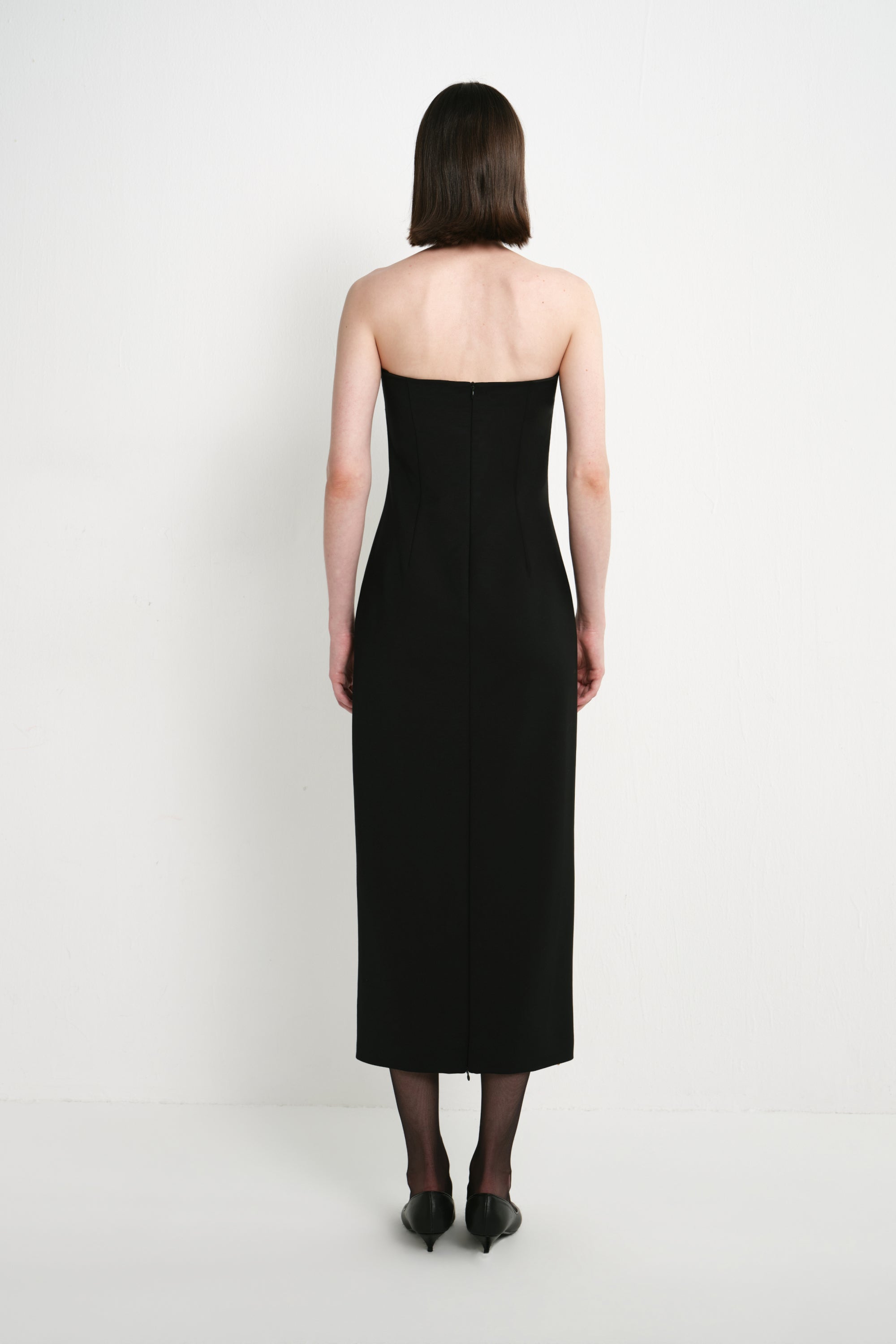 Back view of a model wearing the Rue Sophie Webster Tube Dress in black, highlighting the strapless silhouette, shaping seams, center-back zipper, and straight mid-calf hem.