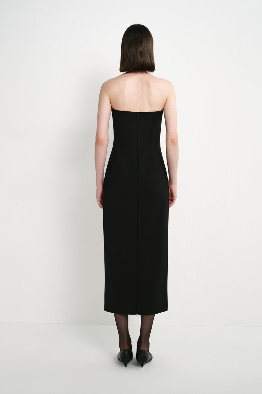 Back view of a model wearing the Rue Sophie Webster Tube Dress in black, highlighting the strapless silhouette, shaping seams, center-back zipper, and straight mid-calf hem.