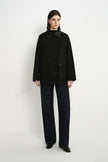 A model stands against a plain white studio background wearing a black button-front jacket with a dark collar and front pockets, paired with dark wide-leg pants and black shoes.