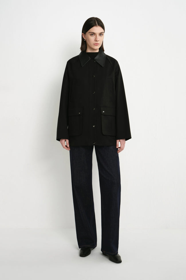 A model stands against a plain white studio background wearing a black button-front jacket with a dark collar and front pockets, paired with dark wide-leg pants and black shoes.