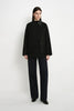 A model stands against a plain white studio background wearing a black button-front jacket with a dark collar and front pockets, paired with dark wide-leg pants and black shoes.