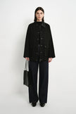 A model stands against a plain white studio background wearing a black button-front jacket over a black top, dark wide-leg pants, and black shoes, while holding a black tote bag.